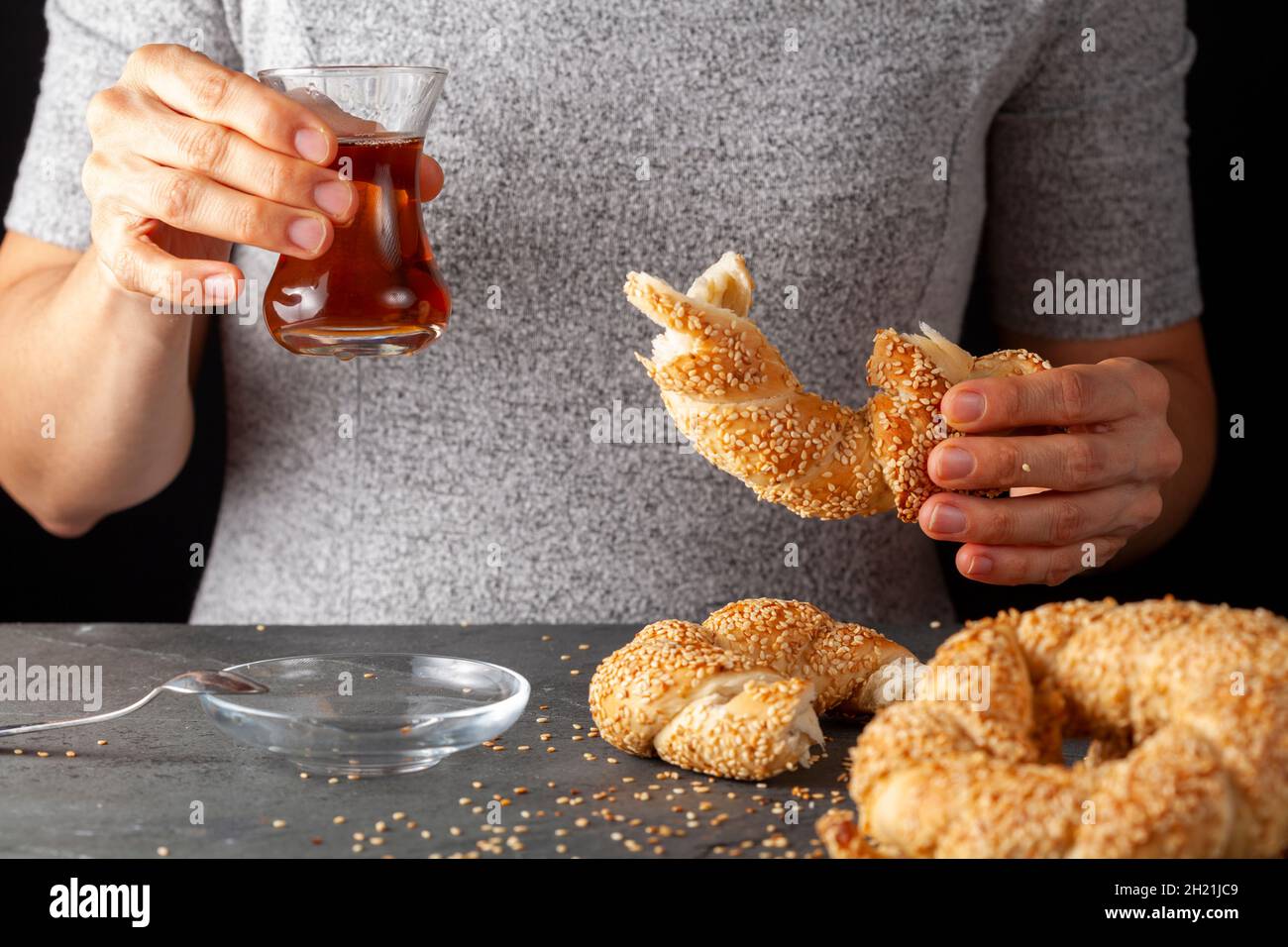 Köstlicher türkischer Bagel mit Sesamsamen, bekannt als Susamli simit. Eine Frau isst ihn zusammen mit schwarzem Tee, der in türkischen Teetassen serviert wird. Dunkle Rückseite Stockfoto