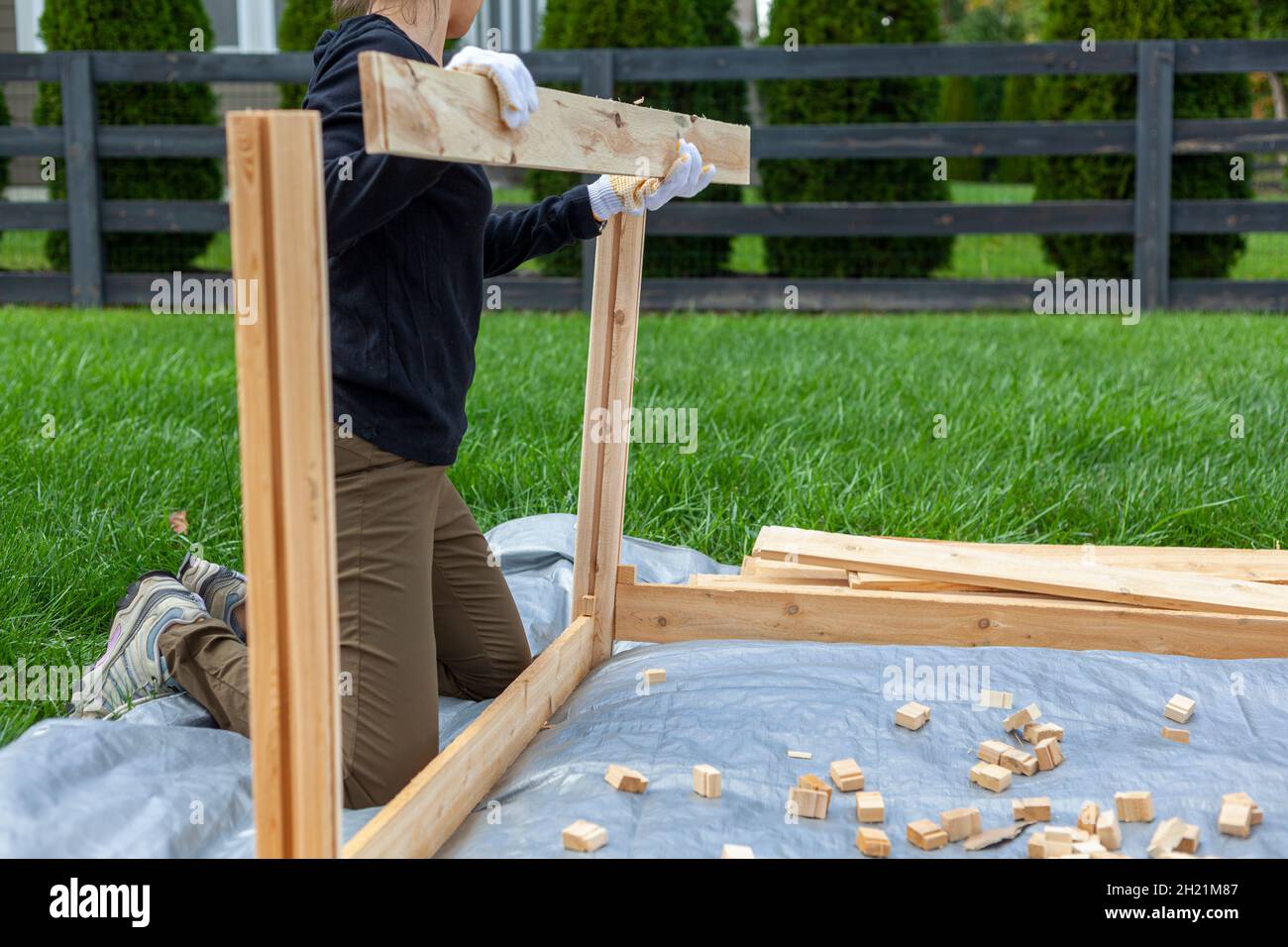 Eine Frau baut einen Holzzaun oder hält sich in ihrem Hinterhof auf Plane. Ein Konzept für die Holzbearbeitung. Möbelinstallation DIY-Projekt. Sie trägt Handschuhe, um p Stockfoto