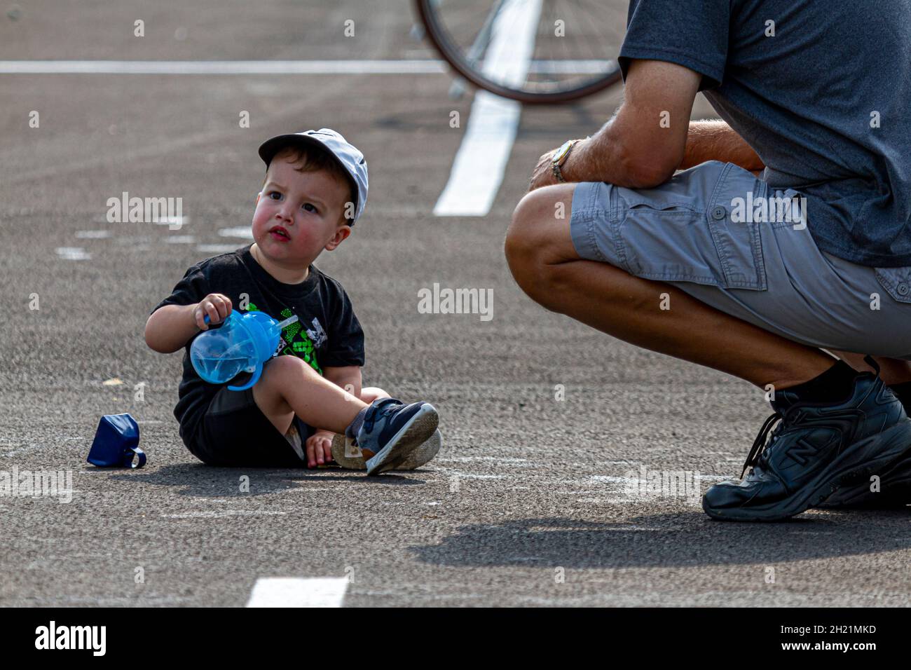 Columbus, OH USA 08-07-2021: Ein süßes blondes Kleinkind sitzt auf Asphalt auf einem Parkplatz und spielt mit einem Spielzeug. Sein Vater kniete neben ihm und p Stockfoto