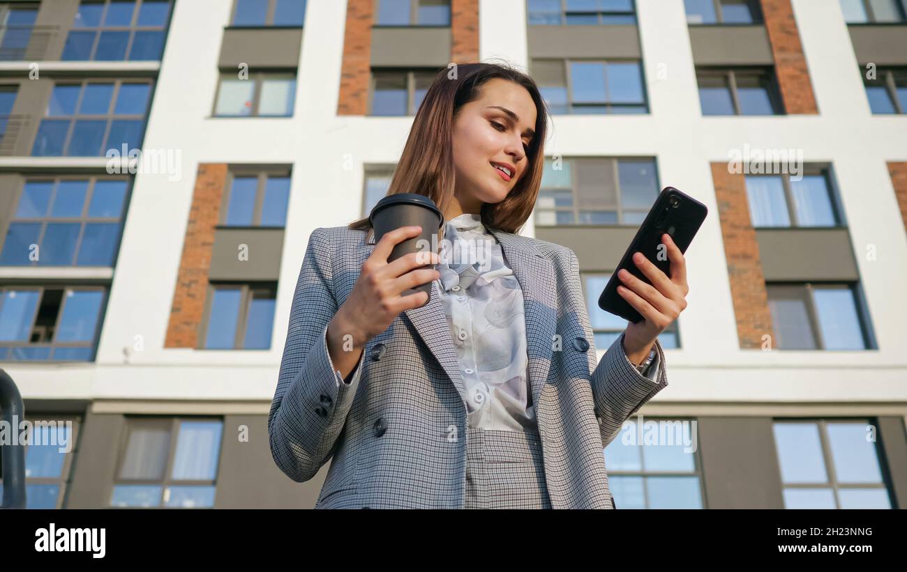 Frau in einem Geschäftsanzug blickt auf das Telefon, während sie eine Plastikschale auf dem Hintergrund eines Gebäudes hält Stockfoto