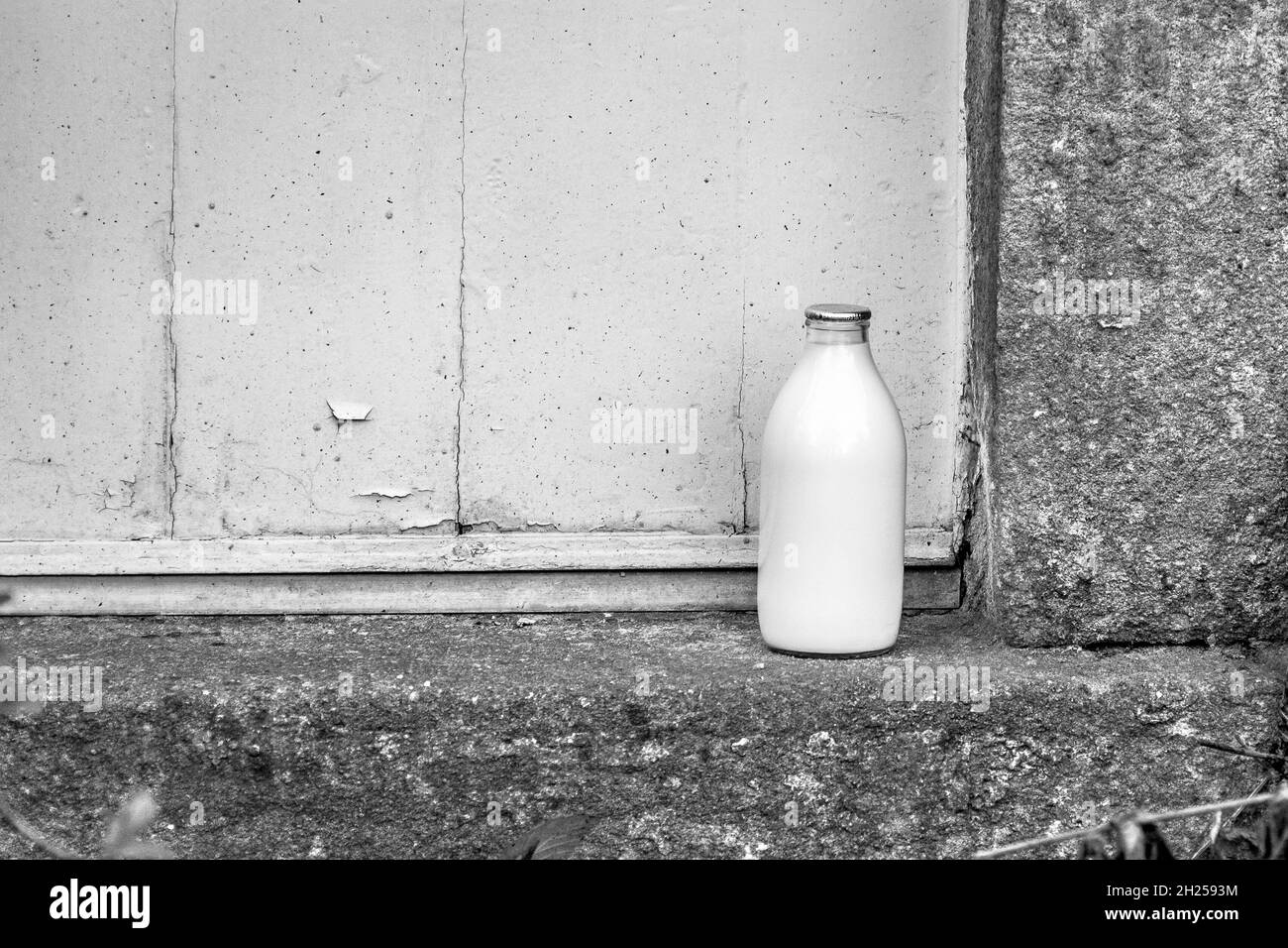 Eine Milchflasche aus Glas mit silberner Oberseite, die mit einer Steinstufe und einer Holztür mit abblätternder Farbe zu einem Haus geliefert wird. Schwarz und Weiß. Stockfoto