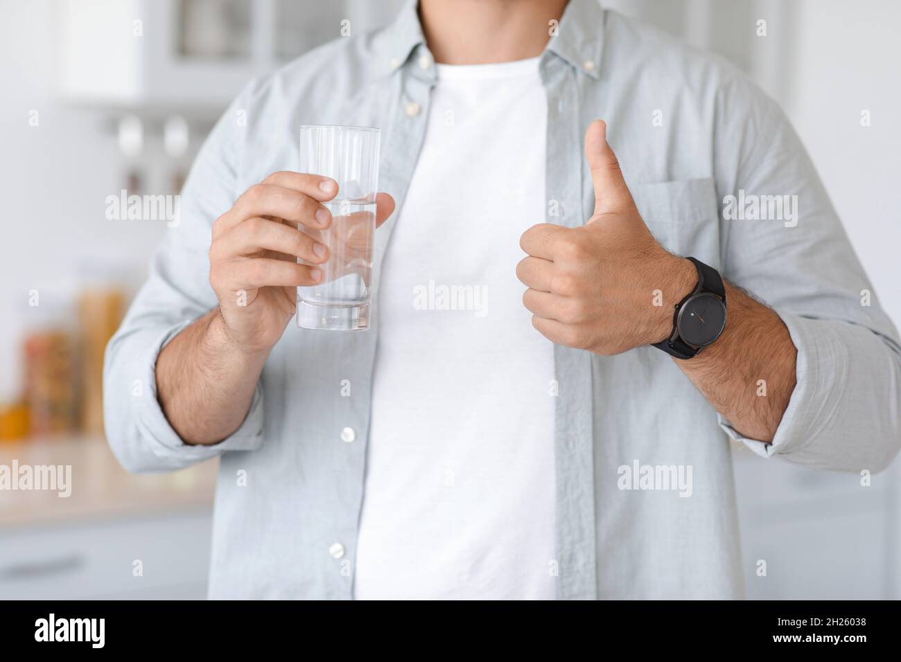 Unkenntlich erwachsener kaukasischer Mann mit einem Glas Wasser, das auf der minimalistischen Kücheneinrichtung ein Daumenschild zeigt Stockfoto