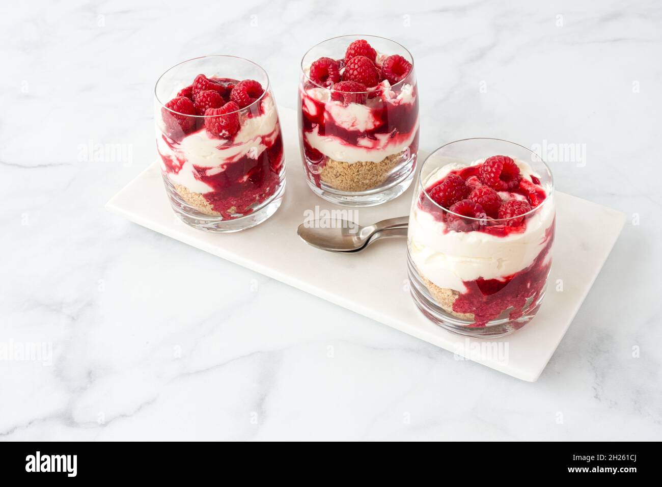 Drei Gläser gefüllt mit hausgemachtem Himbeer-Dessert auf weißem Marmorbrett und weißem Marmorhintergrund mit viel Platz zum Kopieren. High-Angle-Ansicht. Stockfoto
