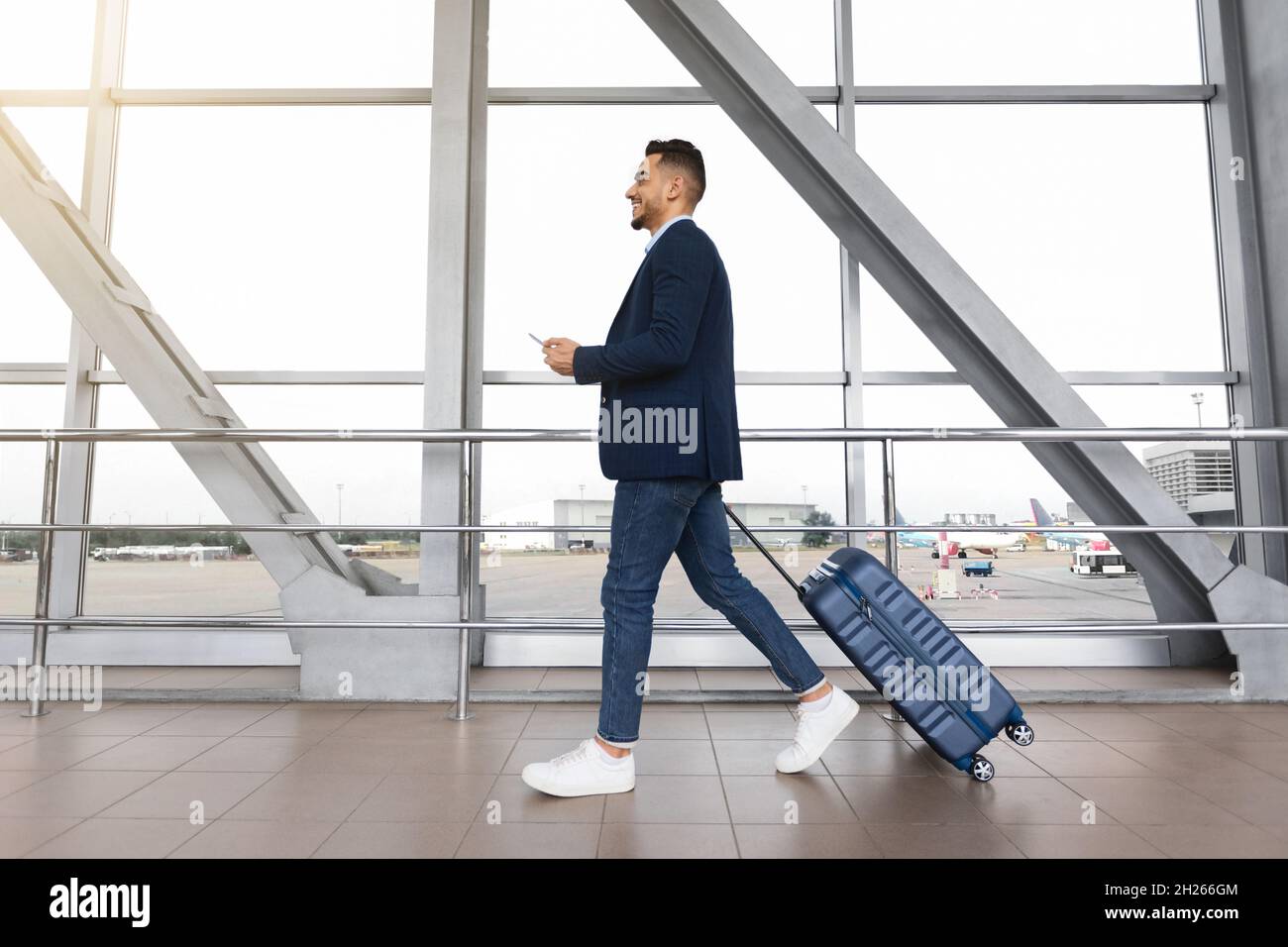 Junger Geschäftsmann aus dem Nahen Osten mit Koffer und Smartphone in den Händen, der auf dem Flughafen unterwegs ist Stockfoto