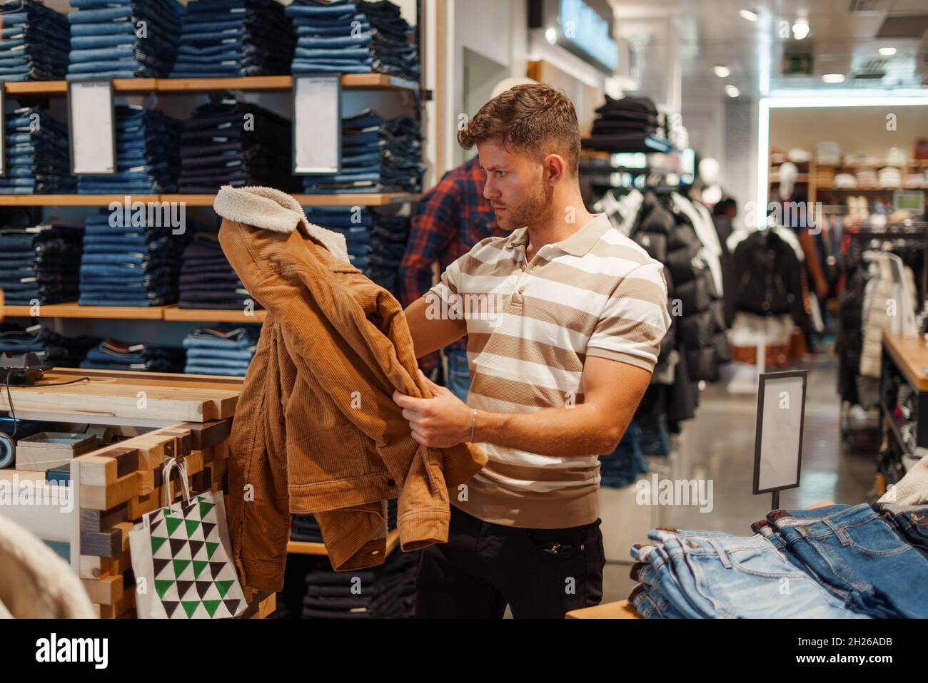 Männlicher Kunde im gestreiften T-Shirt, der die stylische warme Jacke beim Einkaufen in der Kleiderabteilung des Einkaufszentrums anfasst und inspiziert Stockfoto