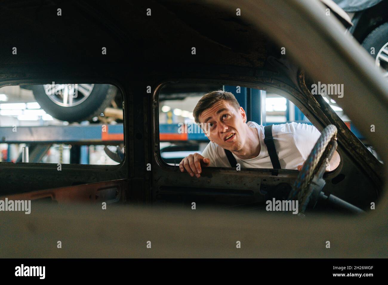 Vorderansicht des frustrierten jungen Service-Mannes in Uniform, der das Innere des alten Autos in der Autowerkstatt mit Fahrzeughintergrund inspiziert. Stockfoto