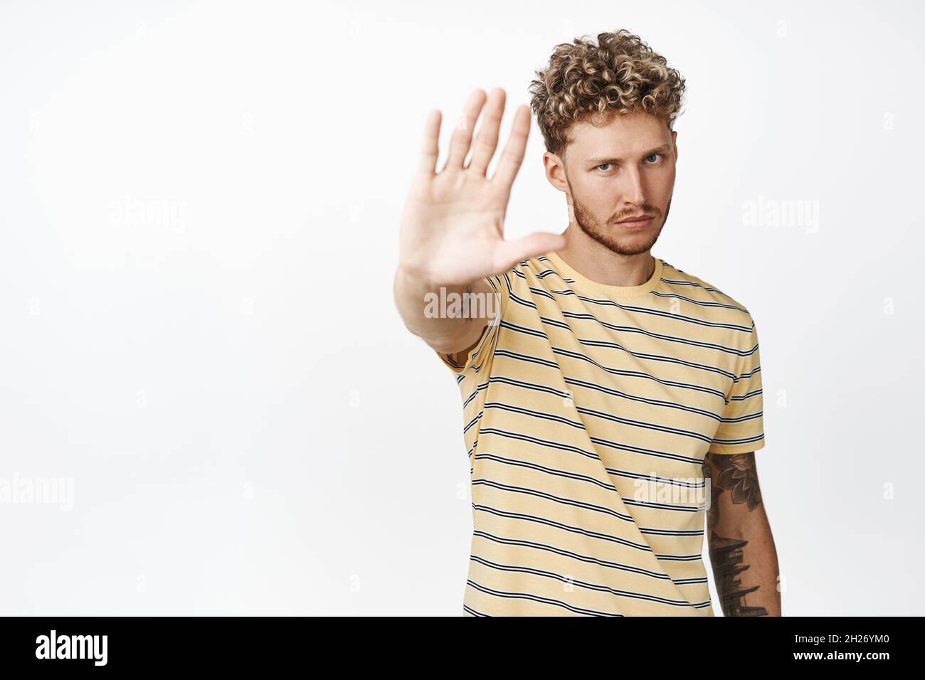 Seriöse blonde lockige Mann Strecke die Hand aus und sieht selbstbewusst aus, sagt zu stoppen, nein zu sagen, zu verbieten, im T-Shirt auf weißem Hintergrund zu stehen Stockfoto