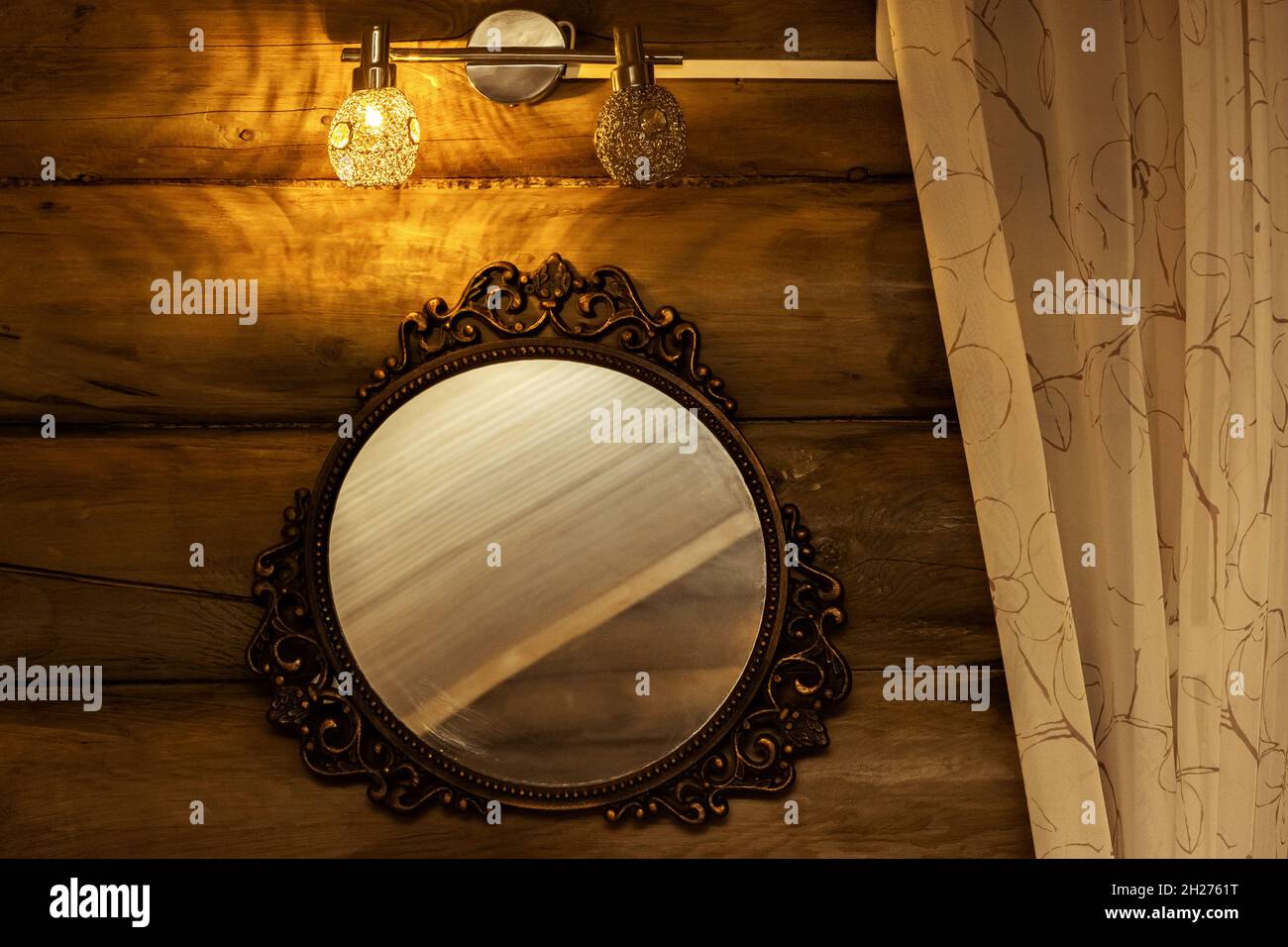 An einer Holzwand hängt ein dekorativ gerahmter runder Spiegel unter einer Lampe neben einem Vorhang. Stockfoto