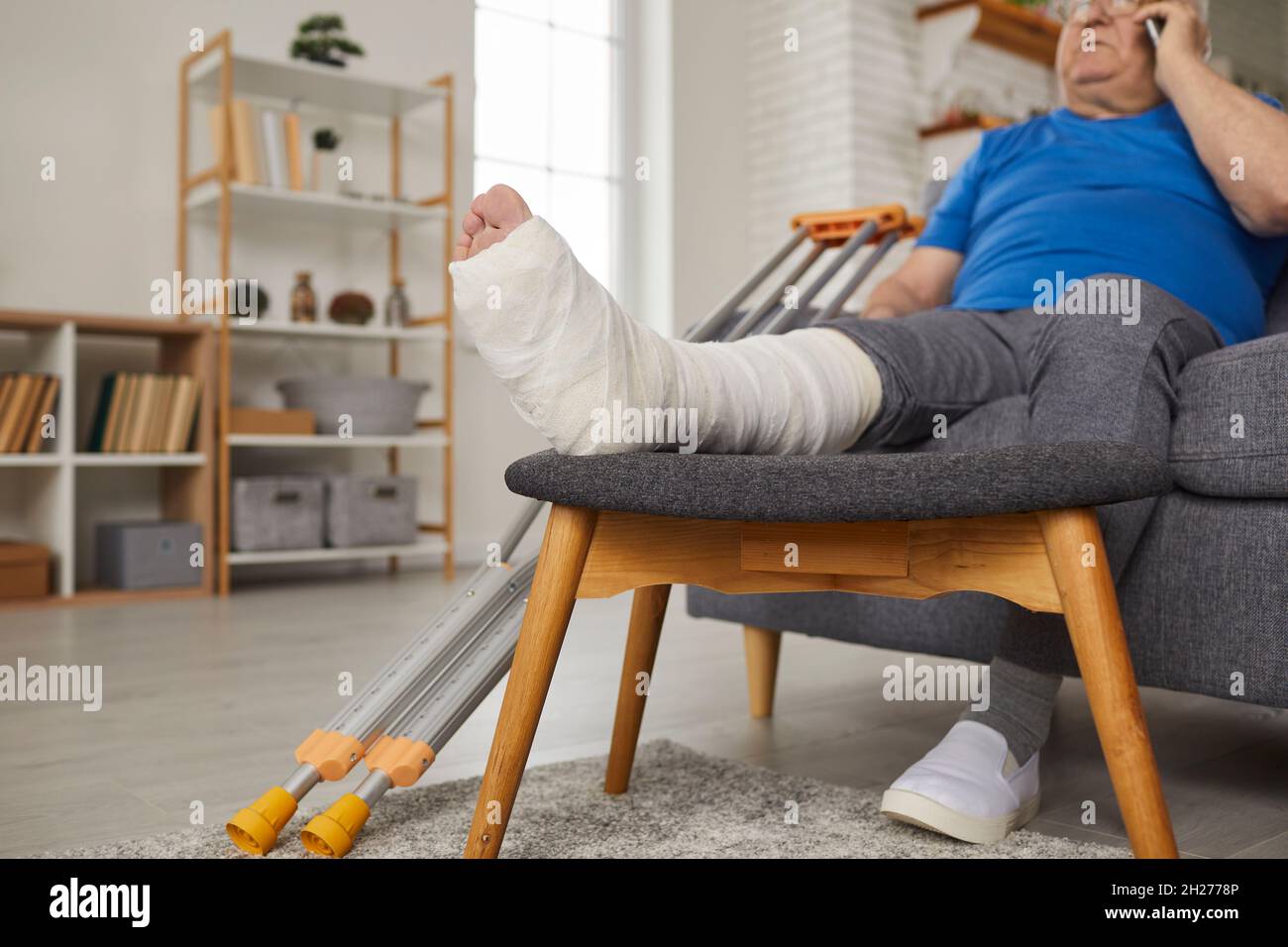 Älterer Mann mit gebrochenem Bein in Gipsabguss sitzt auf der Couch und spricht am Telefon Stockfoto