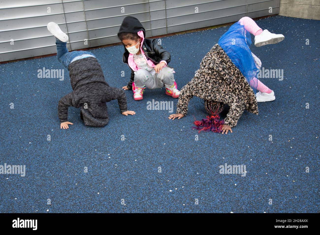 Bildung Vorschule 4-5 Yar alt zwei Mädchen und ein Junge versuchen, Handstände oder Kopf steht draußen auf dem Spielplatz zu tun, tragen Gesichtsmasken Covid-19 Stockfoto