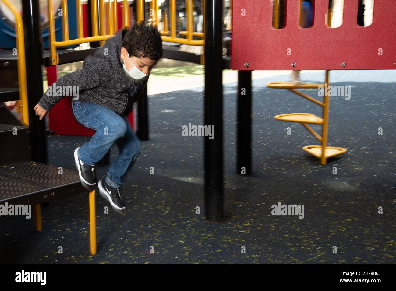 Bildung Vorschule 4-5-Jährige im Freien auf dem Spielplatz Junge springt von Spielgeräten, trägt Gesichtsmaske, um vor Covid-19-Infektion zu schützen Stockfoto