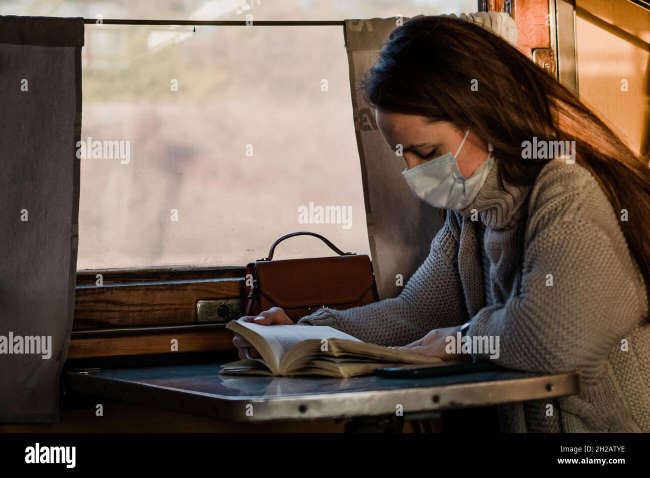 Eine junge Frau in einer medizinischen Maske reist während einer Coronavirus-Pandemie mit dem Zug. Das Mädchen liest ein Buch und hört Musik im Zug. T Stockfoto