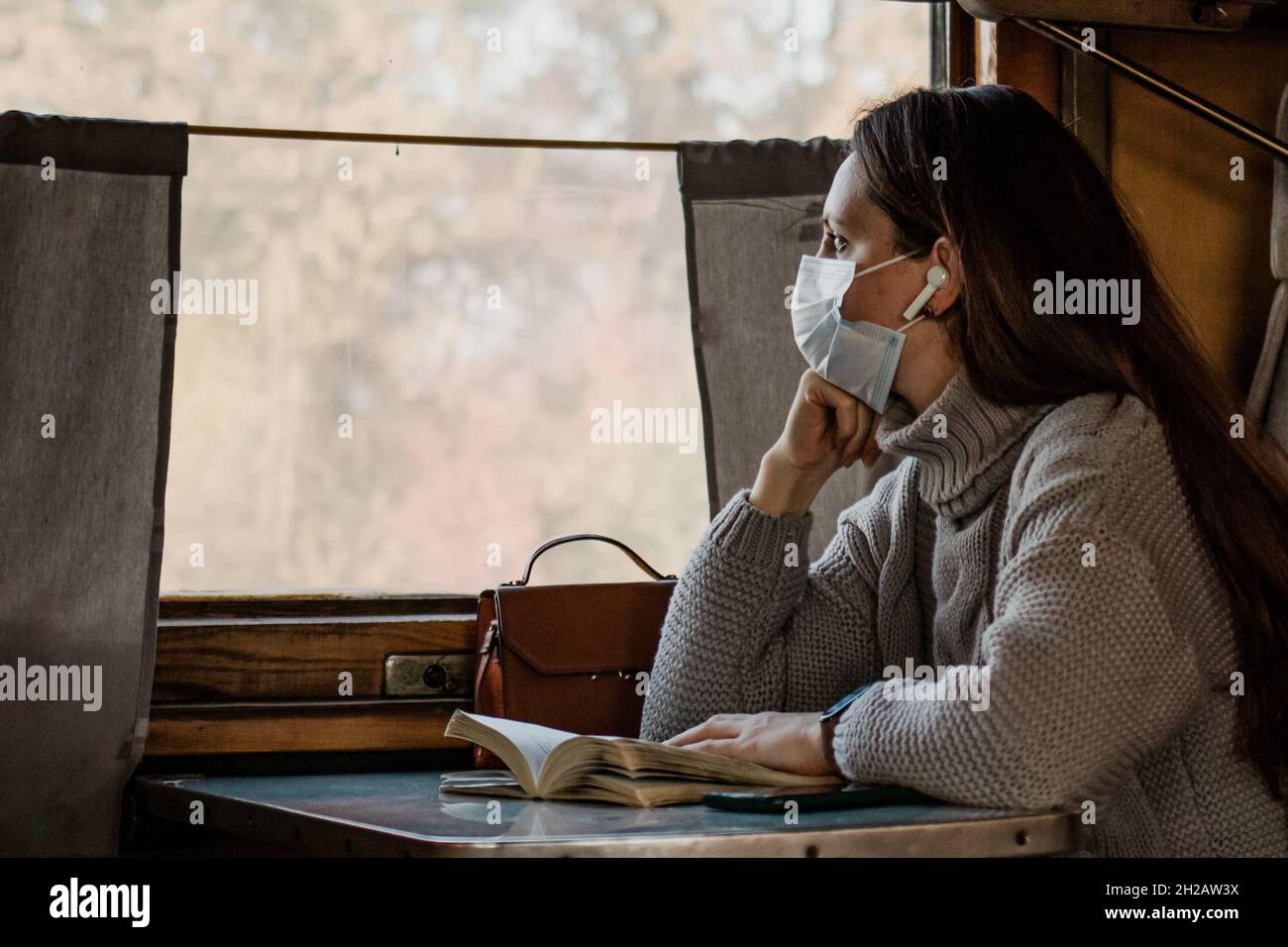 Eine junge Frau in einer medizinischen Maske reist allein mit dem Zug. Das Mädchen schaut ins Fenster und hört Musik im Zug. Einnahme von Preautio Stockfoto