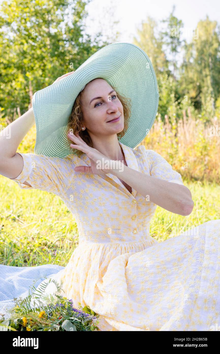 Porträt einer jungen attraktiven kaukasischen Frau in einem leichten, stilvollen Kleid und einem großen Strohhut. Glückliche Dame im Sommerkleid genießt einen Sommertag am Stockfoto