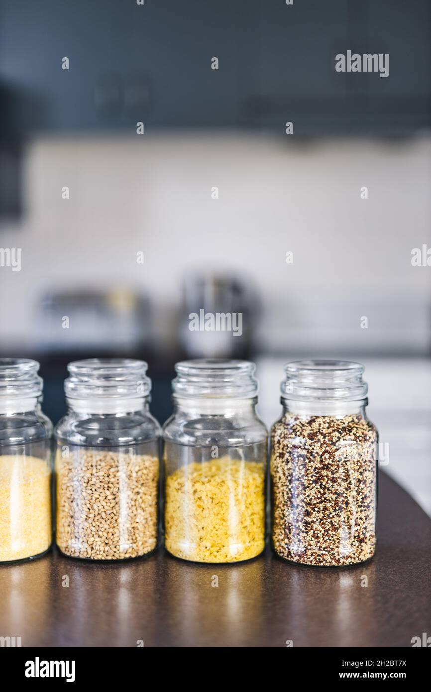 Klare Pantry-Gläser mit verschiedenen Körnern und Hülsenfrüchten, einschließlich Quinoa-Linsen Buchweizen und Gerste auf der Arbeitsfläche mit Küche Bokeh, einfach Ing Stockfoto