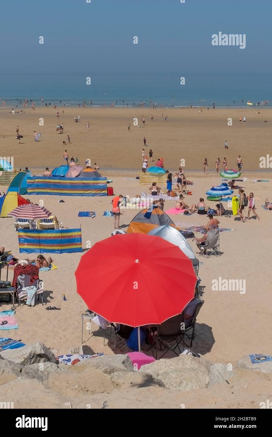 Ein lebhafter roter Sonnenschirm, der von Urlaubern an einem belebten Fistral Beach in den Sommerferien in Newquay in Cornwall verwendet wird. Stockfoto