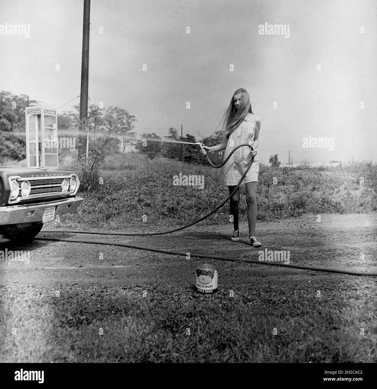 1960er Jahre, historisch, draußen auf einem ländlichen Asphaltweg, ein Teenager-Mädchen, das ein amerikanisches Plymouth-Auto, Virginia, USA, mit einem Jetspray spült. Stockfoto