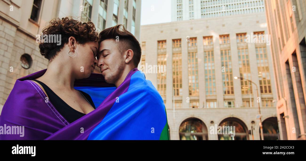 LGBTQ+ Paar steht mit einer Regenbogenfahne um sie herum. Liebevolles junges queeres Paar, das nahe beieinander steht und die Stirn berührt. Ja Stockfoto