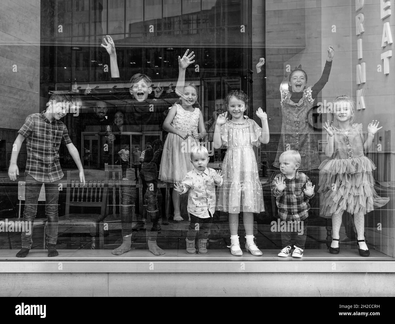 London, Greater London, England, Oktober 09 2021: Eine Gruppe von Kindern lächelt hinter einem Fenster, winkt und tanzt mit einem Roboter. Monochromes Bild. Stockfoto