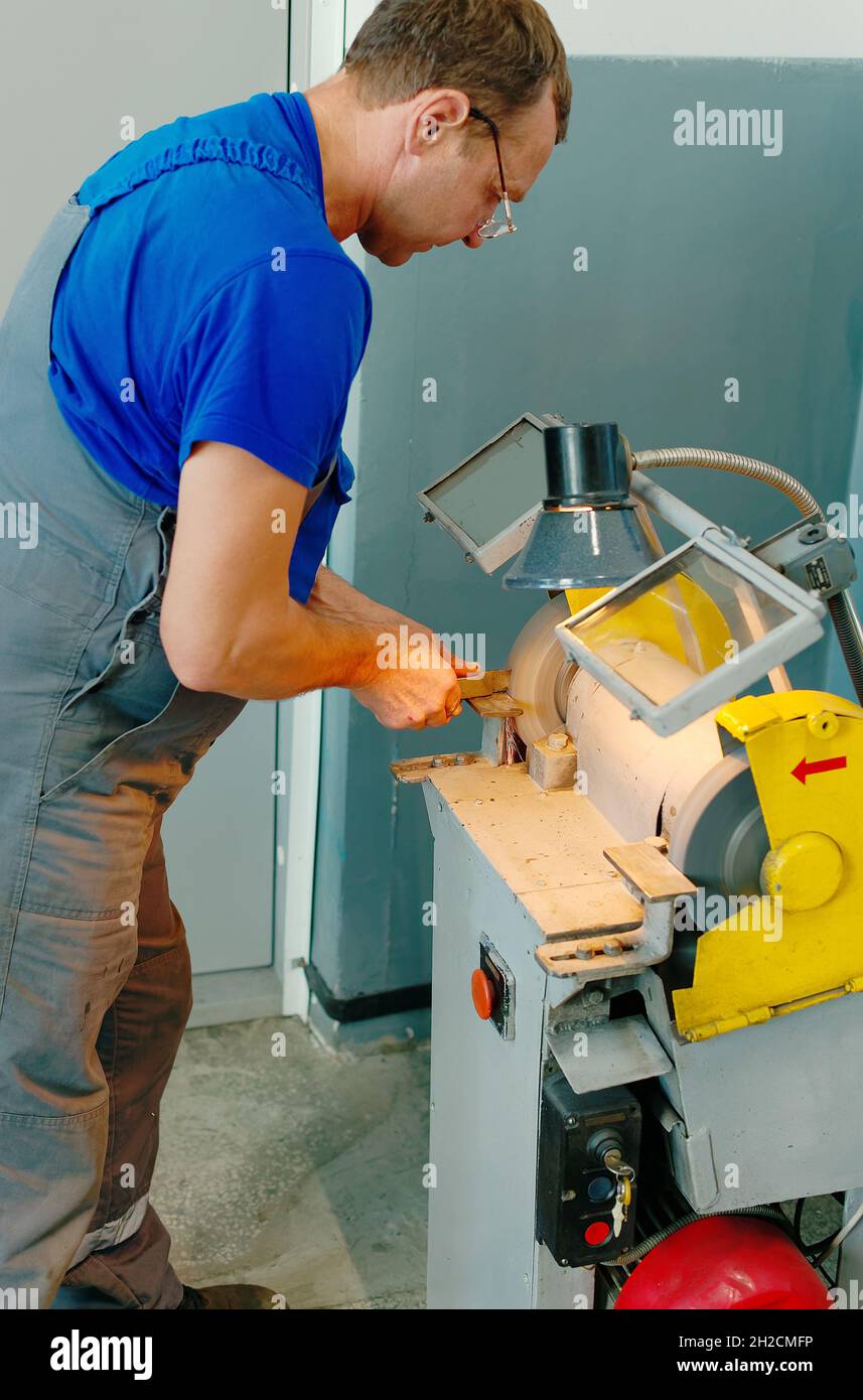 Ein kaukasischer Arbeiter in Uniform schärft in einer Werkstatt ein Teil an einer Maschine. Manuelle Verarbeitung von Metall. Ein wahres Porträt eines Arbeiters. Stockfoto