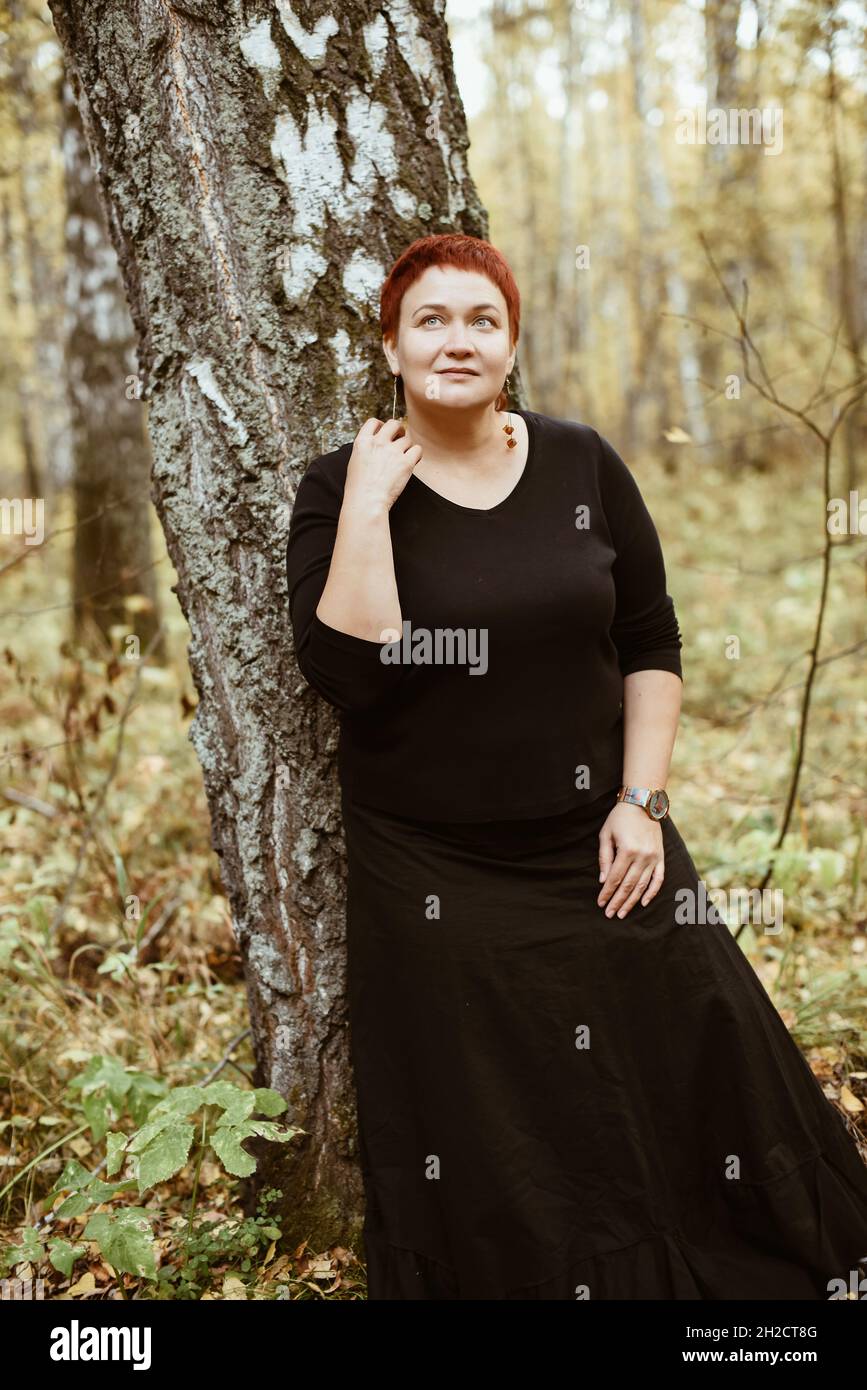 Frau mittleren Alters von 30-40 Jahren im Herbstwald steht am Baum, nachdenklich, verträumt. Stockfoto