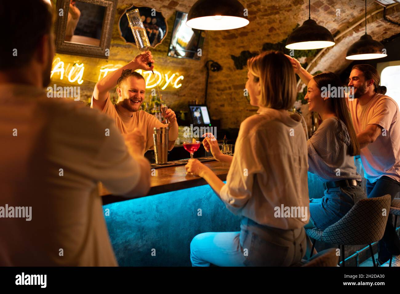 Fröhlicher Barkeeper, der Likör in die Tasse gießt, während er ein Getränk für verschiedene Kunden zubereitete Stockfoto