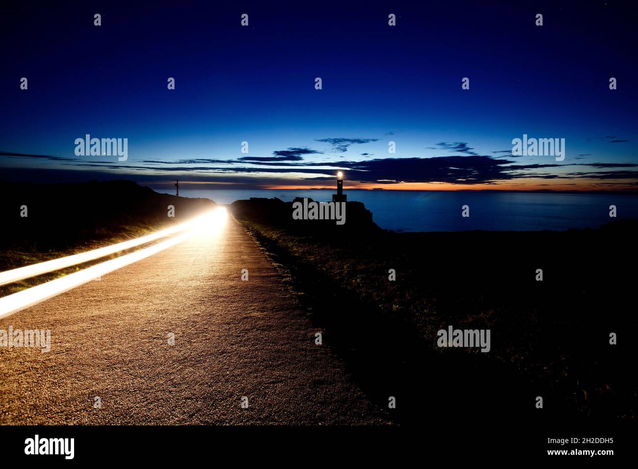 Spur von Autoleuchten am Leuchtturm von Punta Nariga, der sich in der Abenddämmerung an der Küste des Todes im Nordwesten Spaniens befindet Stockfoto