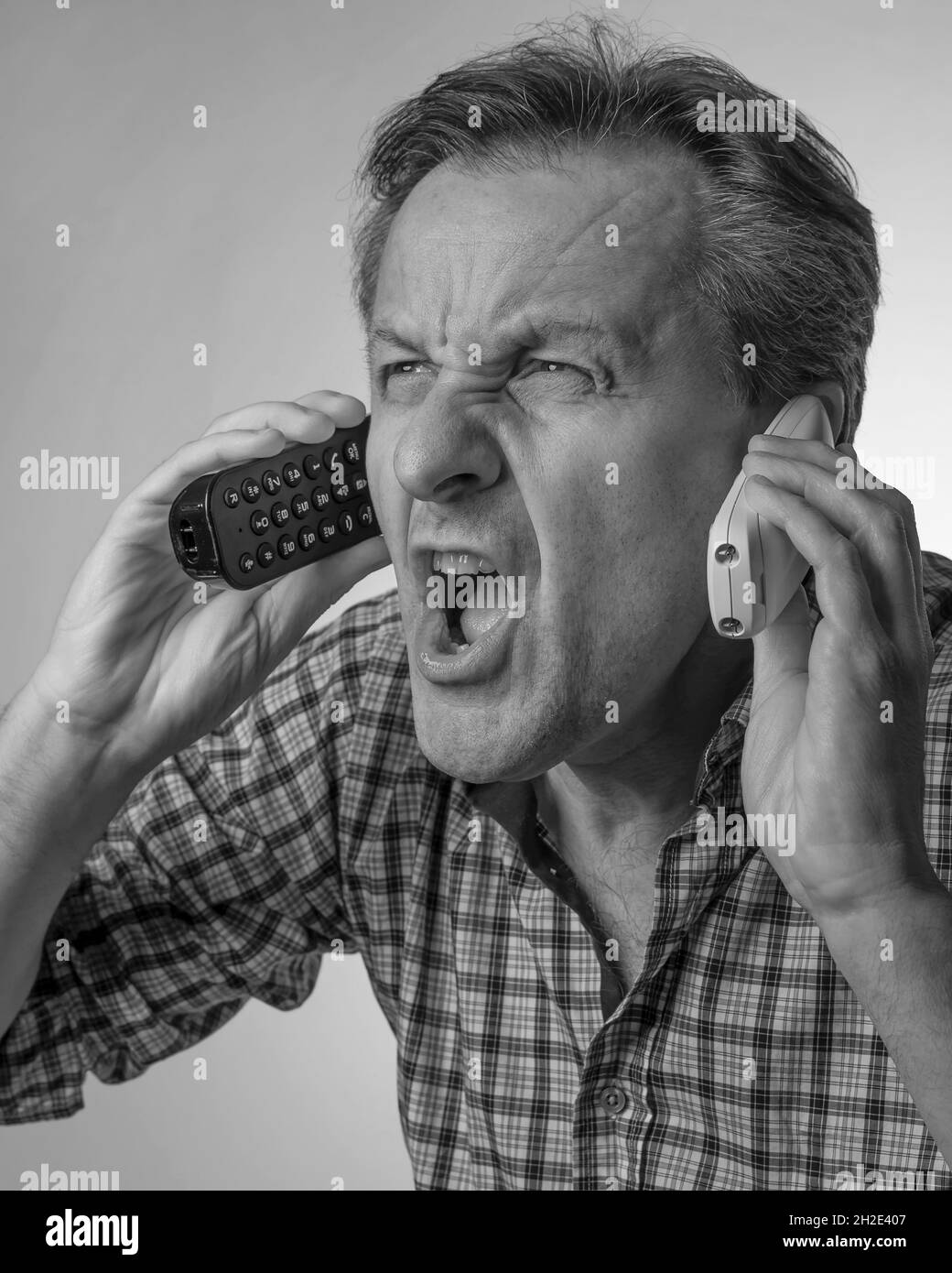 Ein weißer Mann brüllt, während er zwei schnurlose Telefone hält, mit einem Ausdruck von großem Stress, in Schwarz und Weiß Stockfoto