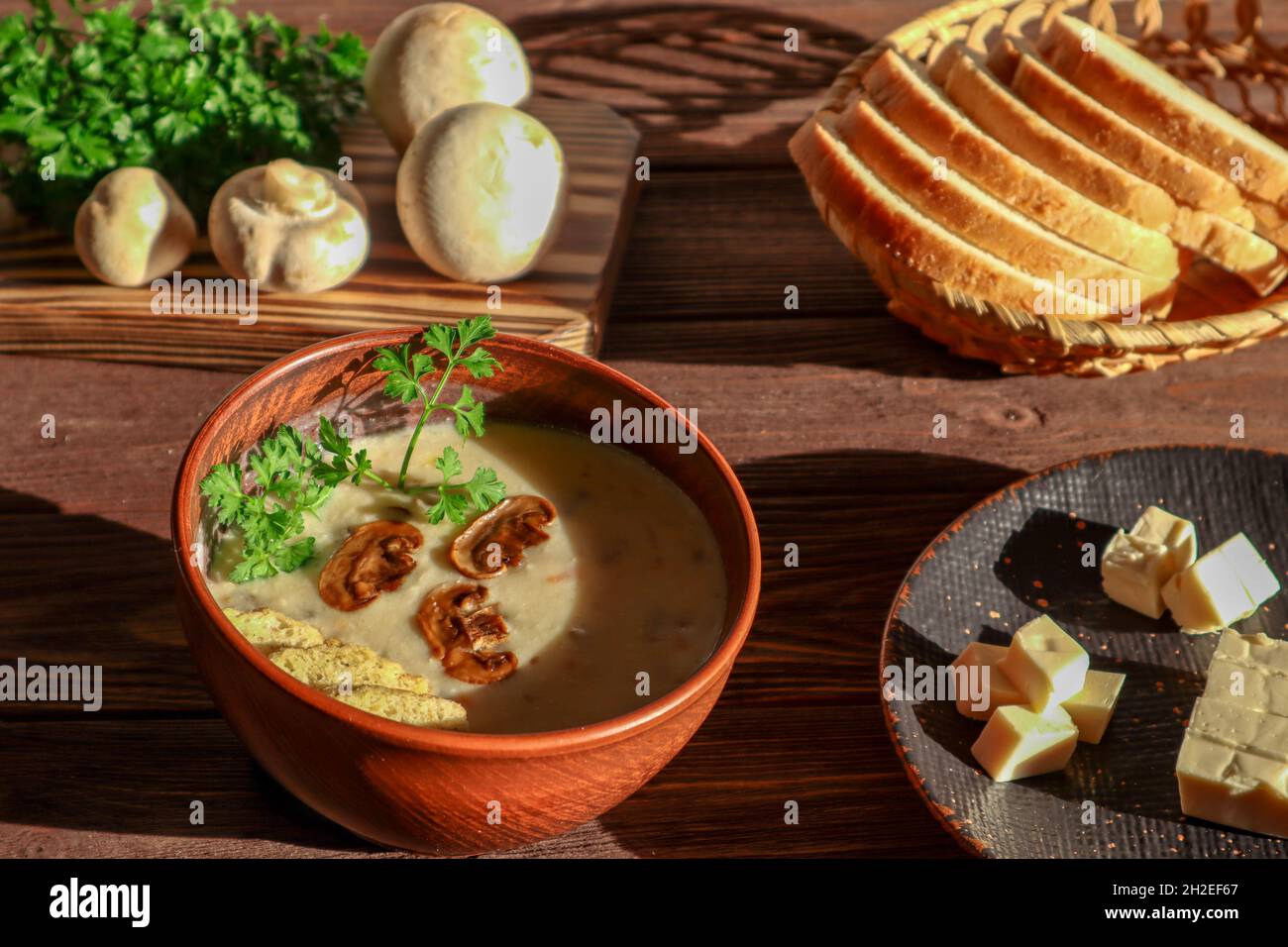Champignon-Suppe mit geschmolzenem Käse, Petersilie und Croutons in einer Schüssel auf einem dunklen Holztisch vor einem Hintergrund von Pilzen und Brot in der hellen Stockfoto