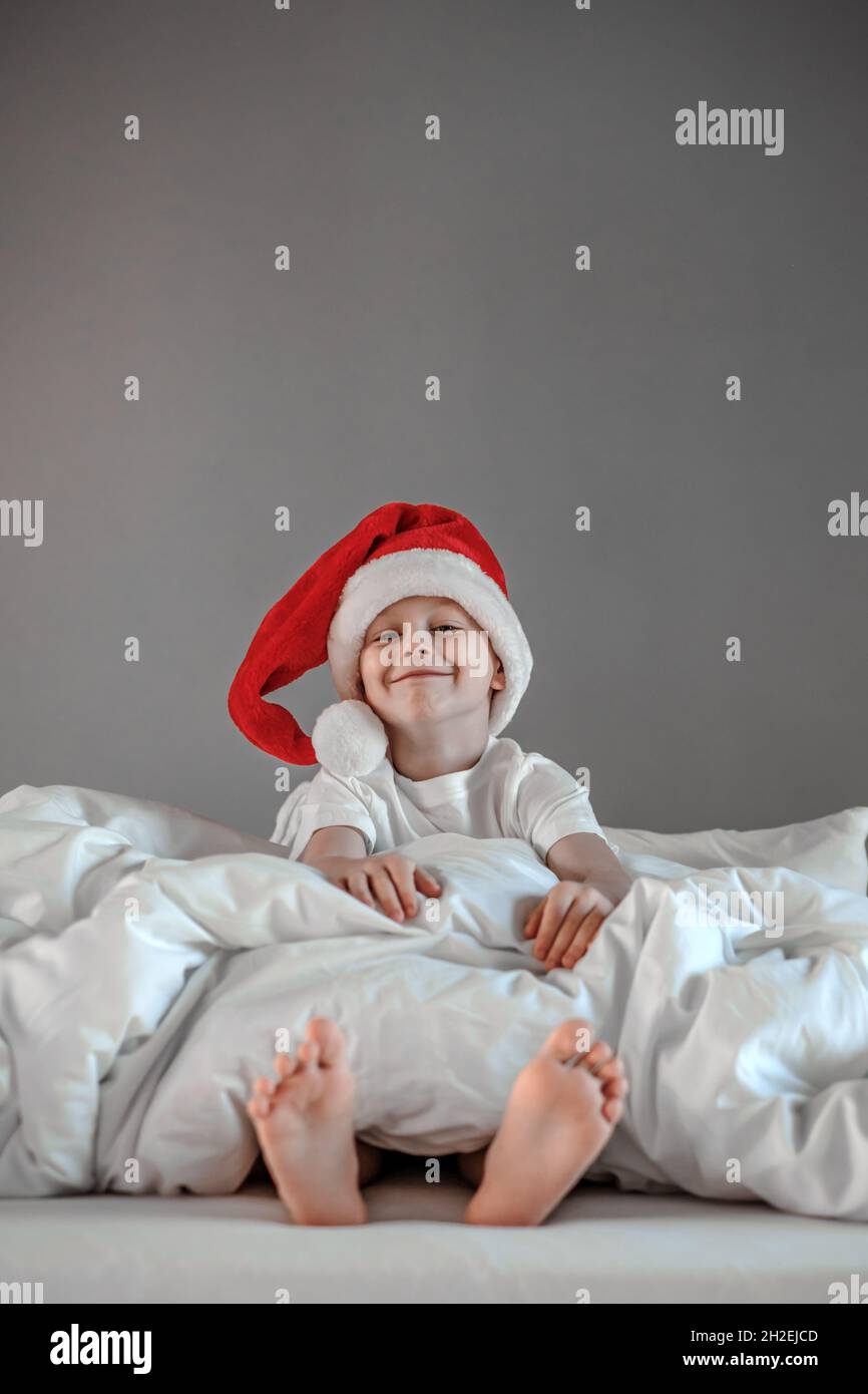 Witziger netter Junge sitzt auf dem Bett und nippt in einem Weihnachtsmütze. Frohe Feiertage. Konzept für den Weihnachtsmorgen Stockfoto