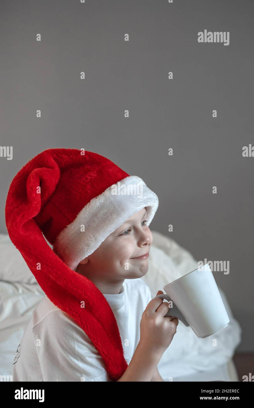 Portrait niedlicher Junge sitzt auf dem Bett mit einem Becher in einem Weihnachtsmütze. Frohe Feiertage. Genießen Sie den Weihnachtsmorgen. Platz für Text Stockfoto