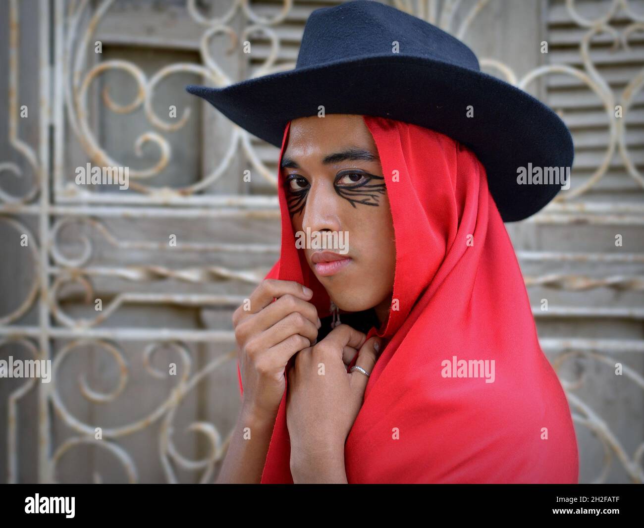 Der hübsche junge, nicht binäre Latino trägt einen schwarzen Hut mit rotem mexikanischem rebozo als Kapuze und posiert vor einem weißen schmiedeeisernen Fenstergrill. Stockfoto