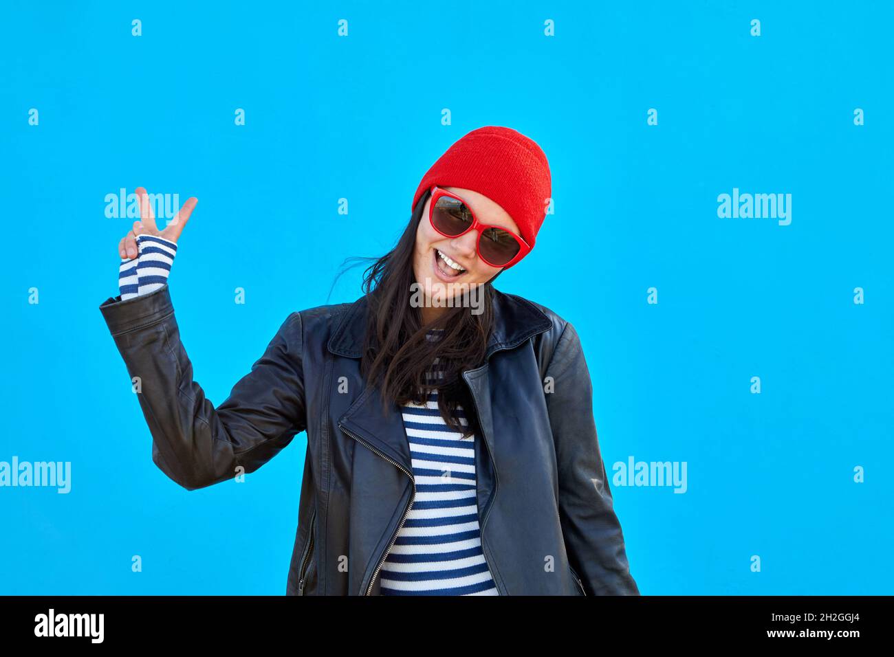 Entzückende trendige Frau in Sonnenbrille und Lederjacke zeigt zwei Finger Geste auf lebhaftem blauen Hintergrund Stockfoto