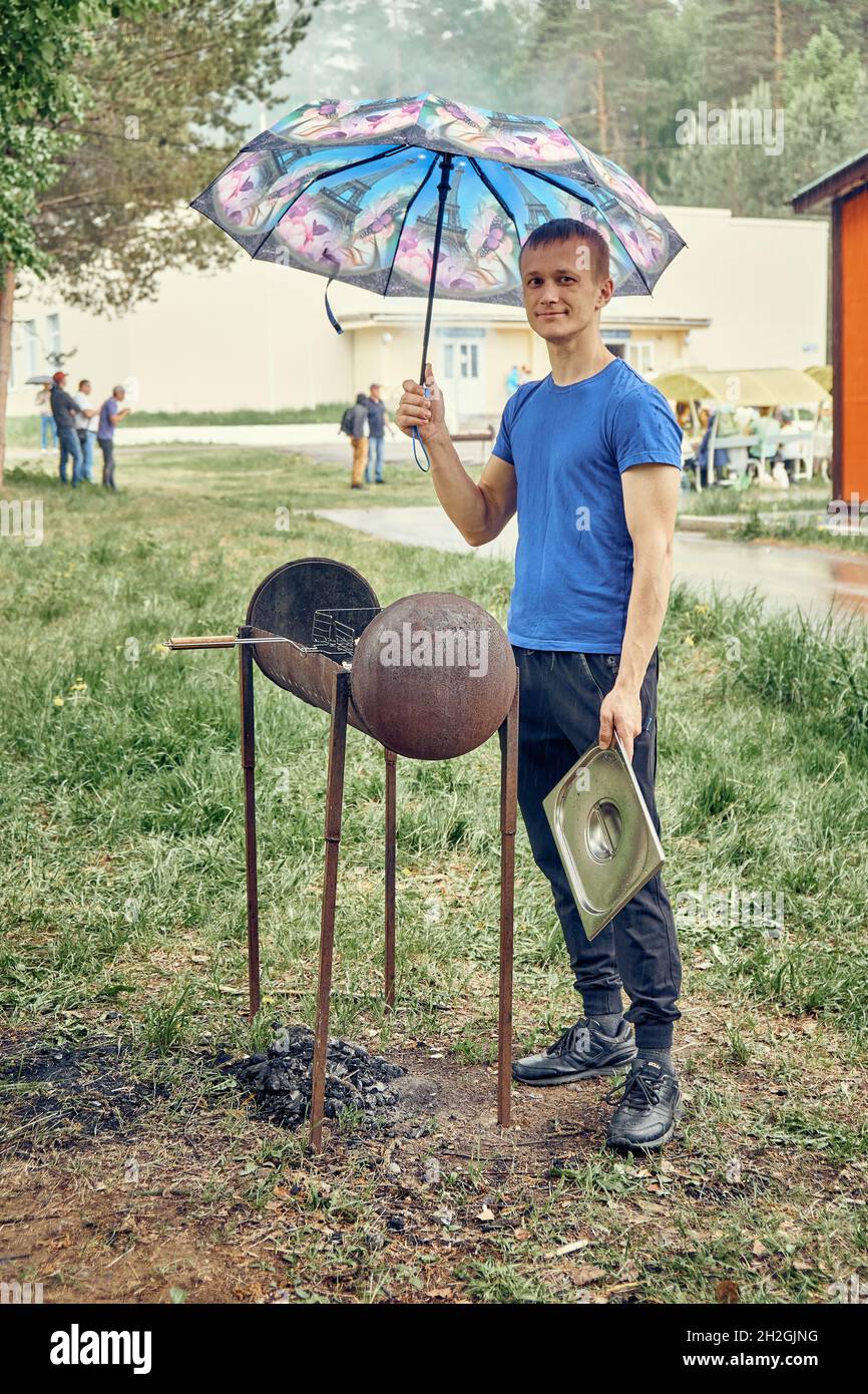 Izhevsk, Russland - 22 2021. Mai: Positiver junger Mann mit Regenschirm kocht Essen auf großem Brazier auf grünem Rasen im öffentlichen Picknick-Stadtpark am regnerischen Frühling Stockfoto