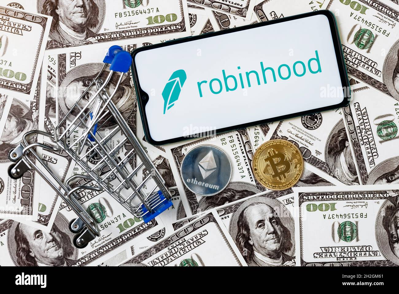 Ein Smartphone mit dem Robinhood-Logo, ein Einkaufswagen und eine Kryptocoins auf den Dollarscheinen. Stockfoto