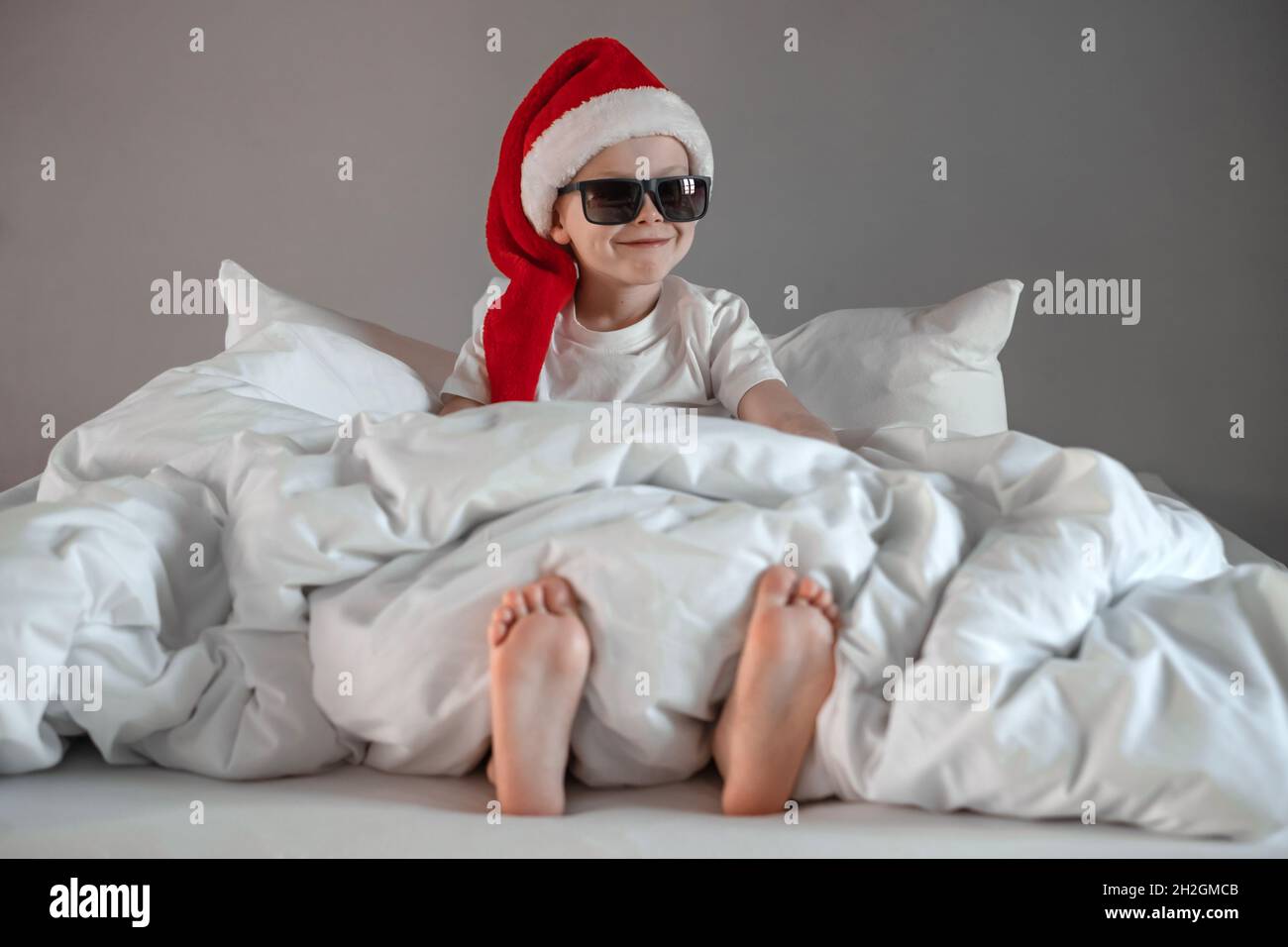 Lustiger, niedlicher Junge sitzt in einer Sonnenbrille auf dem Bett und nippt in einem Weihnachtsmütze. Frohe Feiertage. Konzept für den Weihnachtsmorgen Stockfoto
