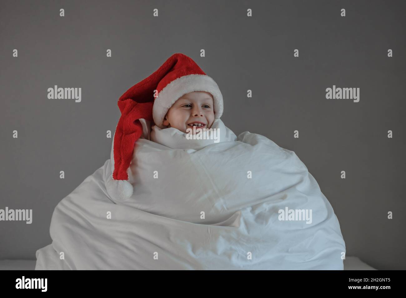 Der siebenjährige Junge wickelte sich in eine weiße Decke in einen Weihnachtsmütze, während er auf dem Bett saß und lächelte Stockfoto