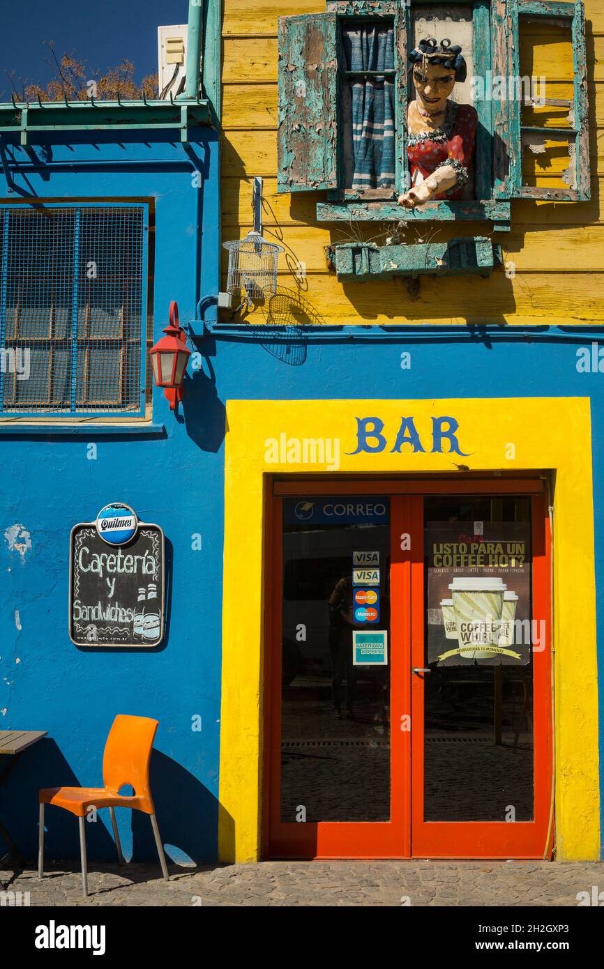 Vertikale Ansicht einer Frauenfigur, die sich aus einem alten Fenster über einen farbenfrohen Bareingang im Viertel La Boca, Buenos Aires, Argentinien, lehnt Stockfoto