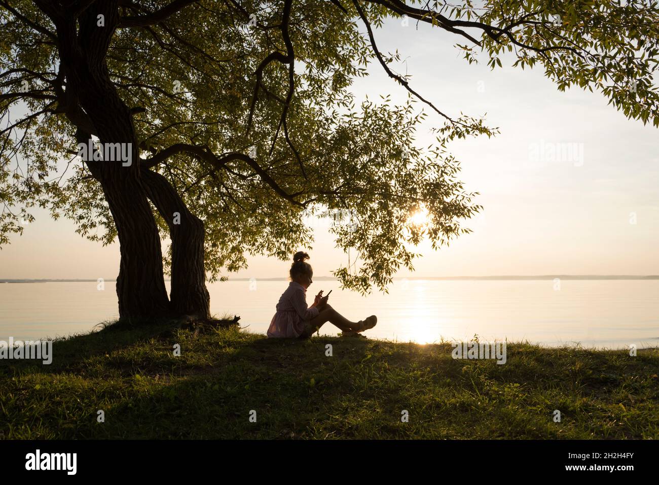 Ein Kind sitzt bei Sonnenuntergang unter einem Baum. Schöne künstlerische Foto mit einem Mädchen in der Natur an einem sonnigen Sommerabend. Träume, Fantasien, Inspiration, Kindheit, Innenwelt und Kinderlebenskonzept. Hochwertige Fotos Stockfoto