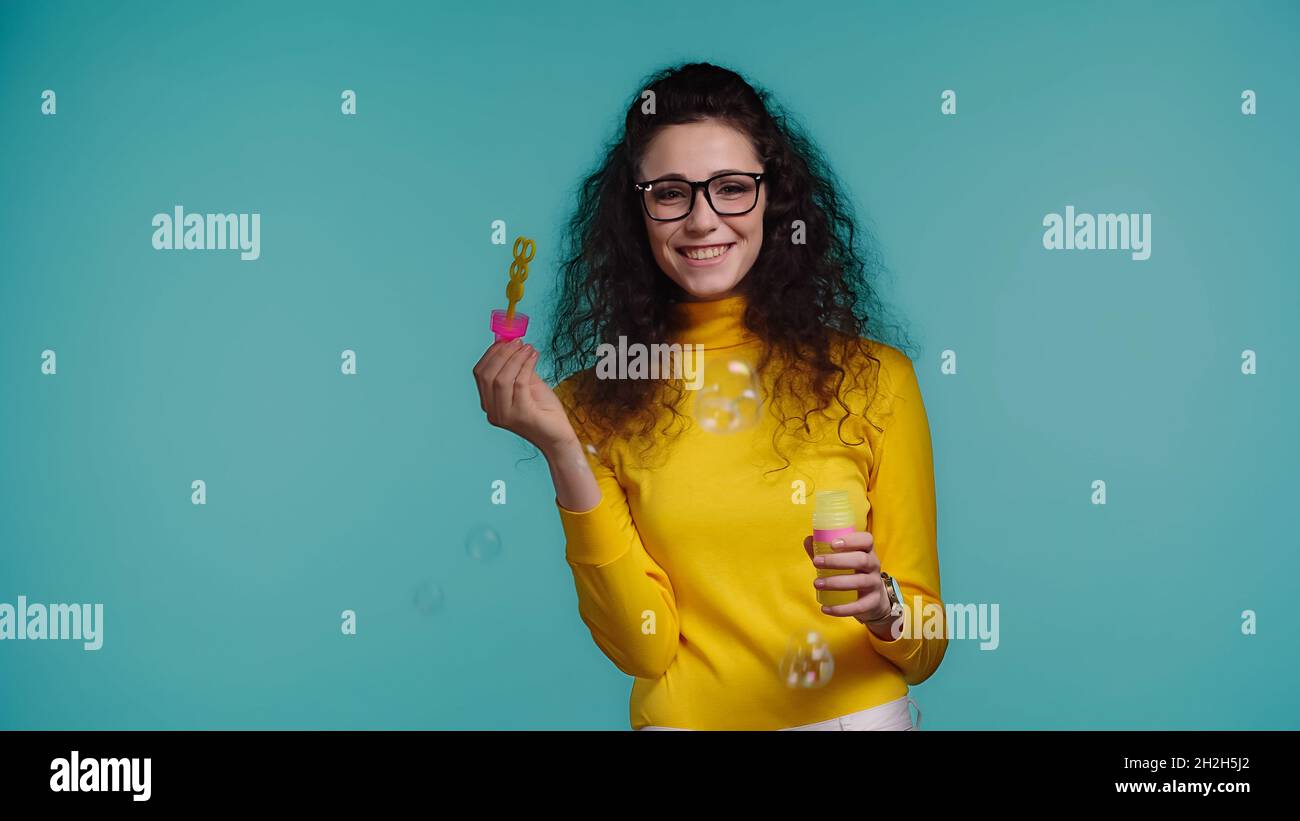 Fröhliche junge Frau in Gläsern mit Flasche mit Seifenblasen isoliert auf blau Stockfoto