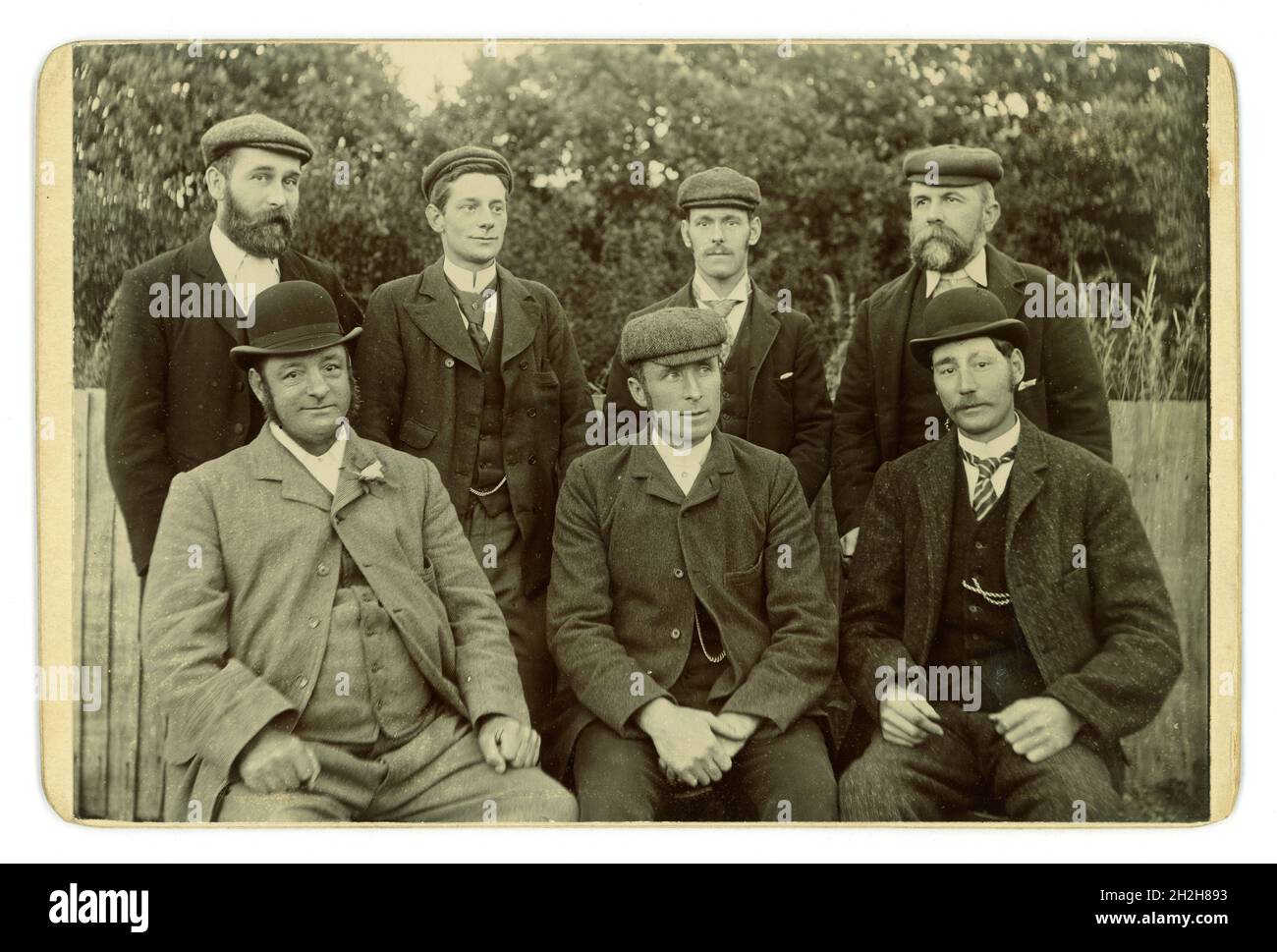 Wunderbare original viktorianische Kabinettkarte einer Gruppe von 7 charaktervollen, glücklich aussehenden, gut gelaunten Herren, verschiedener Altersgruppen, in einer ziemlich informellen und entspannten Pose für die Zeit. Diese Männer sind Mittelklasse, da sie Taschenuhren tragen, die zu der Zeit ein teures Statussymbol waren, und die Fotos zeigen die Männer, die die Uhrenketten mit offenen Anzugjacken zeigen, die draußen in einem Garten sitzen. Es gibt gute Beispiele für Mode. Alle tragen 'Sack-Mäntel' oder 'City-Mäntel' aus Wolle und Tweed, Bowler-Hüte, flache Kappen. Der portly Mann trägt eine Seidenrose Knopfloch. Circa 1885, Großbritannien Stockfoto