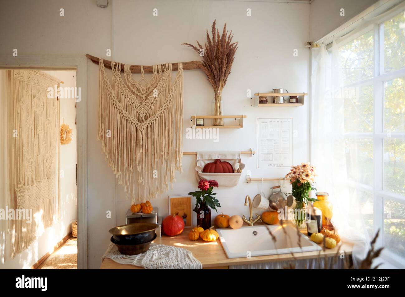 Gemütliche rustikale Küche mit großem Fenster und Spitzenvorhängen. An der Wand hängt ein großes Makrame, Kürbisse, Getreide Stockfoto