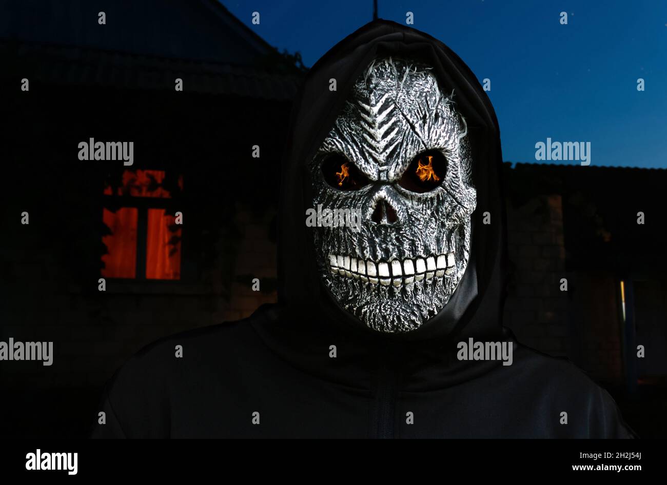 Nahaufnahme des grimmigen Reaper. Mann in Todesmaske mit Feuerflamme in den Augen Nachthimmel, heller Fensterhintergrund. Faschingskostüm, gruselige Zähne. „Hallow“ Stockfoto