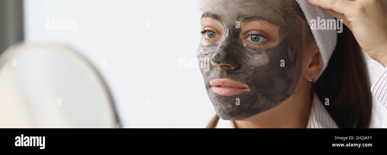 Junge Frau mit schwarzer Tonmaske, die zu Hause in den Spiegel schaut Stockfoto