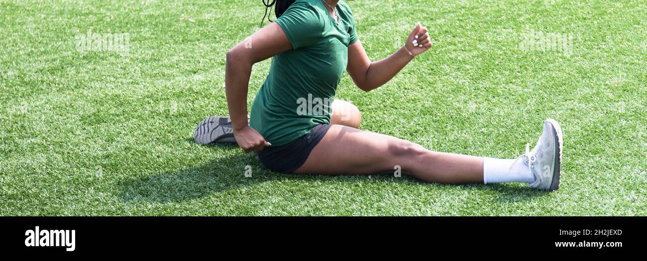 Eine athletische Frau, die ihre Oberschenkelmuskeln aktiv dehnt und dabei eine Streckroutine für Hürden auf einem grünen Rasenfeld durchführt. Stockfoto