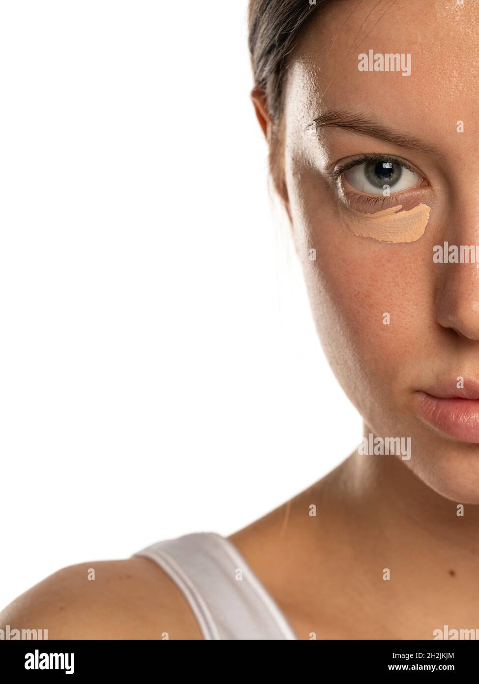 Nahaufnahme einer jungen Frau mit blauen Augen und einem Concealer unter ihrem Auge auf weißem Hintergrund Stockfoto