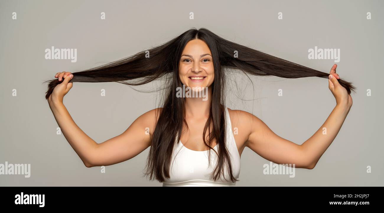 Eine junge glückliche Frau hält und zeigt ihr langes Haar auf grauem Hintergrund Stockfoto