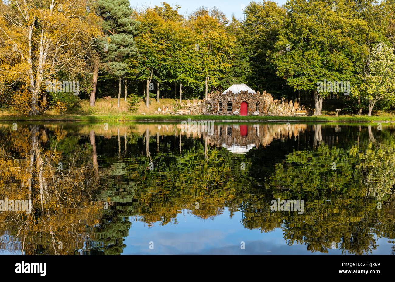 Gosford Estate, East Lothian, Schottland, Großbritannien, 22. Oktober 2021. UK Wetter: Herbstfarben: Sonnenschein beleuchtet Reflexe der Herbstbäume und der schrulligen Steincurling Lodge am künstlichen See im Anwesen Stockfoto