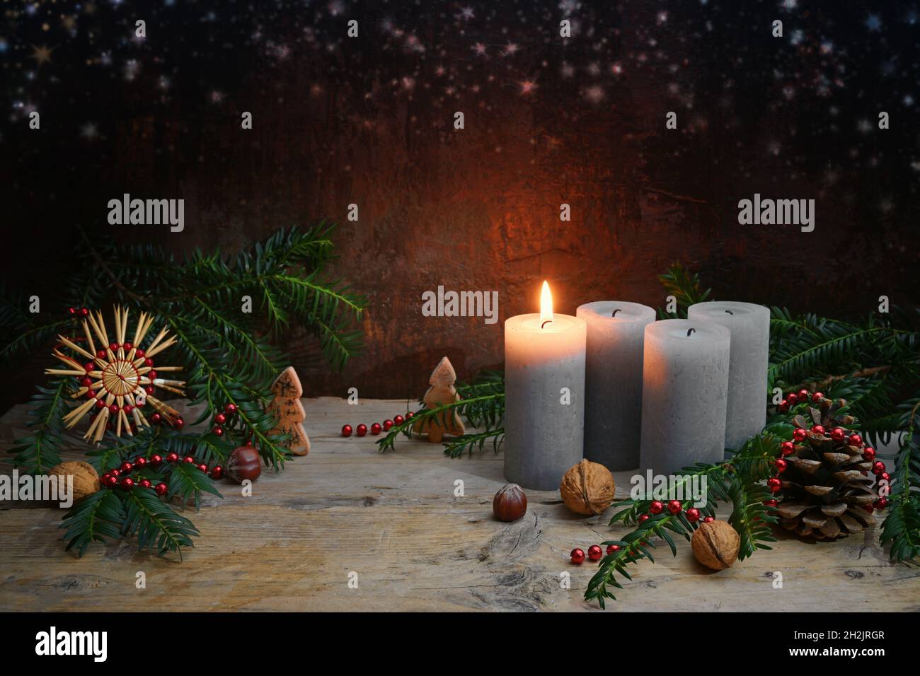 Beim ersten Advent wird eine von vier Kerzen angezündet, Weihnachtsschmuck wie Nüsse, Strohstern, Zapfen und Tannenzweigen auf rustikalem Holz gegen ein dunkelbraunes b Stockfoto