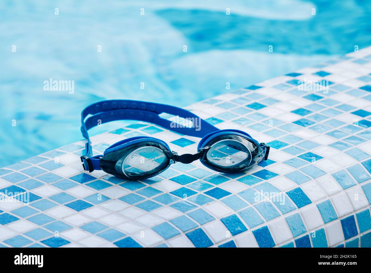 Blaue Profi-Schwimmbrille mit Wassertropfen auf gelben Gläsern auf blauen und weißen Fliesen des Poolbodens. Stockfoto